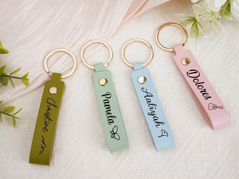 May include: Four personalized keychains in olive green, mint, light blue, and pink leather. Each keychain has a gold ring and a name printed in black script. The pink keychain has a guitar graphic.