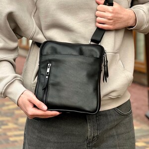 May include: A black leather crossbody bag with a rectangular shape. The bag features a front zipped pocket and an adjustable strap. The bag is held by a person wearing a beige hoodie and grey jeans.