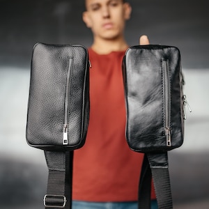 Puede incluir: Dos bandoleras de cuero negro con cremalleras plateadas y correas negras ajustables. Las bolsas tienen forma rectangular con una superficie texturizada. Las bolsas se sostienen frente a una persona que lleva una camisa roja y vaqueros azules.