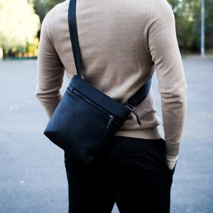 May include: A black leather crossbody bag with a zipper closure and adjustable strap. The bag is worn over the shoulder. The person is wearing a tan sweater and black pants.