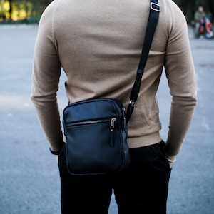 Puede incluir: Un bolso bandolera de cuero negro con cremallera plateada y correa negra ajustable. El bolso se lleva sobre un suéter color beige y pantalones negros. La persona lleva gafas de sol.