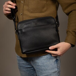 May include: A black leather crossbody bag with a textured surface. The bag has a rectangular shape with a zipper closure and a long, adjustable strap. The bag is worn over the shoulder.