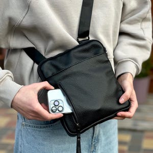 Puede incluir: Bolso bandolera negro con superficie texturizada y bolsillo con cremallera. El bolso tiene una correa larga y ajustable. Un smartphone es parcialmente visible en el bolsillo con cremallera.