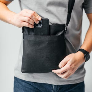 May include: Black leather crossbody bag with a textured finish. The bag has a front pocket and a black strap. A black smartphone is partially visible in the front pocket.