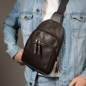 Puede incluir: Un bolso bandolera de cuero marrón oscuro con una superficie texturizada. El bolso tiene dos compartimentos con cremallera con cremalleras doradas y una correa negra ajustable. El bolso se lleva cruzado sobre el pecho.