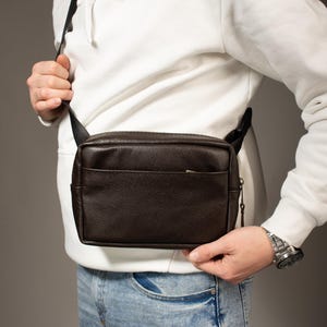 Puede incluir: Bolso bandolera de cuero marrón oscuro de forma rectangular. El bolso tiene un compartimento principal con cremallera y un bolsillo delantero. Se lleva cruzado con una correa negra. La persona lleva una sudadera con capucha blanca y vaqueros azules.
