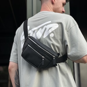Puede incluir: Riñonera de cuero negro con dos compartimentos con cremallera y una correa negra ajustable. La bolsa se lleva cruzada. La persona lleva una camiseta gris con un gráfico blanco en la espalda.