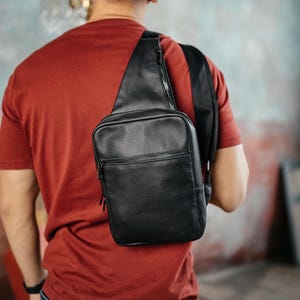 May include: A black leather sling bag with a zippered main compartment and a smaller front pocket. The bag is worn over the shoulder and across the chest. The bag is made of textured leather.