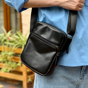 May include: A black rectangular crossbody bag with a zipper closure and adjustable black strap. The bag is made of a smooth, shiny material. The bag is worn over the shoulder.