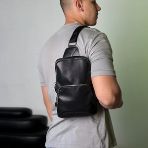 May include: Black leather sling bag with a rectangular main compartment and a smaller front pocket, both with silver zippers. The bag has a wide, adjustable strap and is worn over the shoulder.
