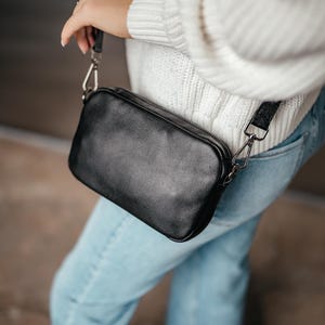 Puede incluir: Un bolso bandolera de cuero negro con forma rectangular y cierre de cremallera. El bolso tiene una correa larga y ajustable y herrajes plateados. El bolso se lleva al hombro. La persona lleva un suéter blanco y vaqueros azules.