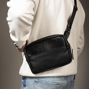 Puede incluir: Bolso bandolera negro con un patrón texturizado similar al de cocodrilo en la parte superior y un acabado de cuero liso en la parte inferior. El bolso se lleva al hombro con una correa negra. La persona lleva una sudadera con capucha blanca y vaqueros azules.
