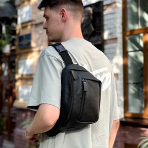 Puede incluir: Un cinturón de cuero negro con un bolsillo frontal con cremallera y una correa ajustable. La bolsa se lleva cruzada. La bolsa está hecha de cuero texturizado y tiene un diseño elegante y moderno. La bolsa se lleva sobre una camiseta gris claro.