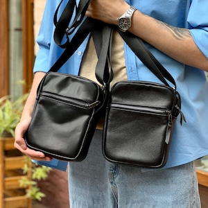 Puede incluir: Dos bolsos negros cruzados con correas negras ajustables. Cada bolso tiene forma rectangular con cierre de cremallera en la parte superior. Los bolsos se sostienen frente a una persona que lleva una camisa azul claro y vaqueros.