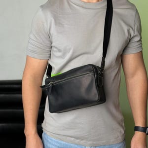 May include: Black leather crossbody bag with a white stitched design and a black adjustable strap. The bag has a zipper closure and a rectangular shape. The bag is worn over the shoulder.