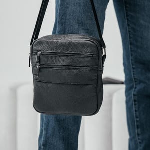 Puede incluir: Bolso bandolera de cuero negro con tres compartimentos con cremallera y una correa larga y ajustable. El bolso es rectangular con una superficie texturizada y se lleva al hombro. La persona lleva vaqueros azules.