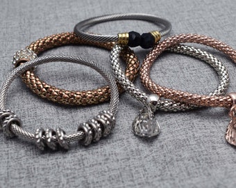 May include: A collection of five bracelets featuring various textures and finishes. Included are silver-tone mesh bands, a twisted cable bangle with black beads, a rose gold-tone mesh bracelet, and a silver-tone bracelet with charms.