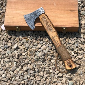 May include: A hand axe with a silver metal head featuring an etched Vegvisir symbol and intricate patterns. The wooden handle has carved details, a wrapped cord grip, and a leather lanyard. The axe rests on a rectangular wooden storage box.
