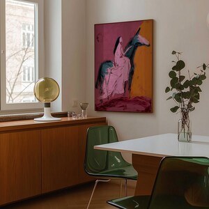 May include: An abstract painting of a person riding a donkey, with pink, black, and orange hues, hangs on a wall. The room features a white table, green chairs, and a wooden cabinet. A decorative lamp and a vase with greenery are also visible.