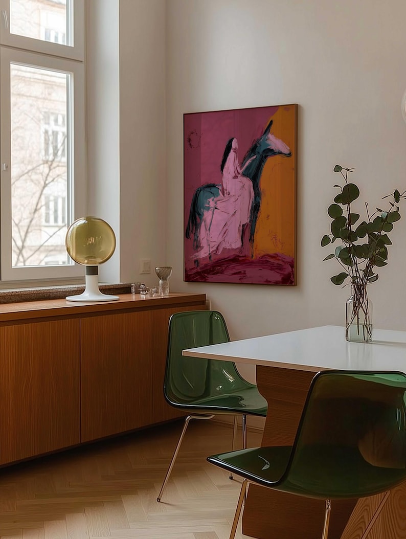 May include: An abstract painting of a person riding a donkey, with pink, black, and orange hues, hangs on a wall. The room features a white table, green chairs, and a wooden cabinet. A decorative lamp and a vase with greenery are also visible.