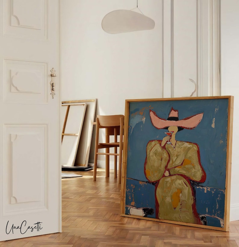 K&ouml;nnte beinhalten: Abstrakte Malerei in einem Holzrahmen, die eine Figur mit gro&szlig;em rosa Hut und gelbem Mantel vor blauem Hintergrund zeigt. Das Kunstwerk wird in einem Raum mit Holzboden und wei&szlig;en W&auml;nden ausgestellt.
