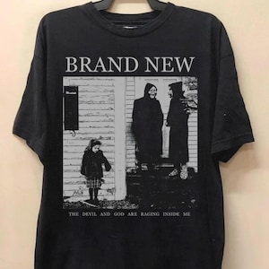 Könnte beinhalten: Schwarzes T-Shirt mit dem Bandnamen "BRAND NEW" über einem Schwarz-Weiß-Bild von drei Figuren. Der Text "THE DEVIL AND GOD ARE RAGING INSIDE ME" ist darunter gedruckt. Das T-Shirt hängt an einem schwarzen Kleiderbügel.