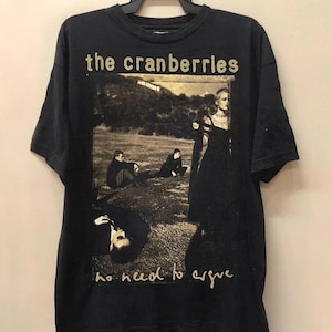 Può includere: T-shirt nera con il nome della band "the cranberries" in lettere dorate sopra un'immagine color seppia. L'immagine raffigura persone in un campo. Il testo "no need to argue" è sotto l'immagine.