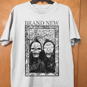May include: A light gray t-shirt featuring the text "BRAND NEW" above a black and white graphic. The graphic depicts two stylized faces, one a skull, within a decorative frame. The shirt has short sleeves.