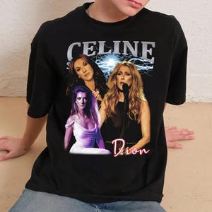 May include: Black t-shirt featuring a graphic of Celine Dion with the text "CELINE" above the images and "Dion" in a neon-style font below. The graphic includes three images of the singer with a lightning bolt background.