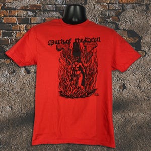 Puede incluir: Camiseta roja con un gráfico negro de una figura en llamas, con el texto "Mark of the Devil" arriba. El diseño está centrado, con una estética vintage.