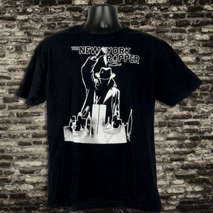 Może przedstawiać: Czarny t-shirt z białym nadrukiem przedstawiającym postać w płaszczu i kapeluszu, podnoszącą mikrofon. Tekst "THE NEW YORK RAPPER" znajduje się nad postacią. Projekt zawiera sylwetkę miasta.