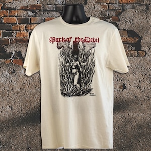 Può includere: T-shirt color crema con un'illustrazione in bianco e nero di una persona in fiamme. Il testo rosso "Mark of the Devil" è stampato sopra. La maglietta è esposta su un manichino.