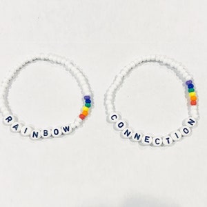 May include: Two white beaded bracelets with rainbow accents. One bracelet spells "RAINBOW" and the other spells "CONNECTION".
