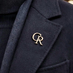May include: A gold-colored lapel pin in the shape of the letters "CR" is affixed to a dark navy blue blazer. The pin is positioned on the lapel, near the buttonhole. The blazer has a textured appearance and is paired with a dark turtleneck.
