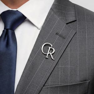 May include: A silver-colored lapel pin with the intertwined letters "CR" is affixed to a gray pinstripe suit jacket. The jacket is paired with a white dress shirt and a navy blue tie, creating a classic, formal look.