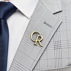 May include: A gold-colored lapel pin in the shape of the letters "CR" is affixed to a gray plaid blazer. The blazer is paired with a white dress shirt and a navy blue tie. The pin adds a touch of elegance to the formal attire.