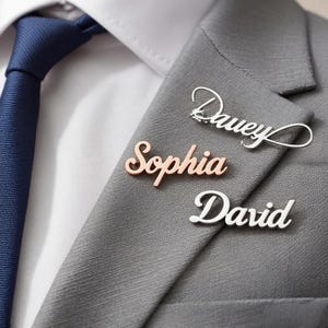 Könnte beinhalten: Nahaufnahme von drei Namensschildern: "Davey" in Silber, "Sophia" in Roségold und "David" in Silber. Die Anstecknadeln sind an einem grauen Anzug befestigt. Eine blaue Krawatte und ein weißes Hemd sind im Hintergrund zu sehen.