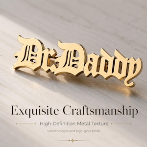 May include: A gold-toned metal brooch featuring the text Dr.Daddy in a stylized gothic font. The piece has a high-gloss finish with smooth, reflective edges. The text is connected as a single unit, showcasing a polished metallic surface.