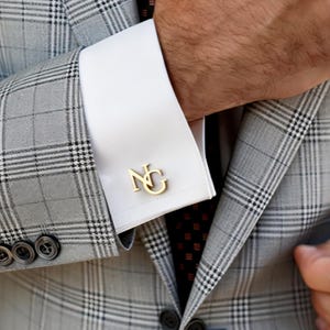 May include: Gold-colored cufflinks with the initials "NG" are displayed on a white dress shirt cuff. The shirt is paired with a gray plaid suit jacket. The image is a close-up of the cuff and jacket.