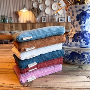 Op de afbeelding: Een stapel van zes rechthoekige, geribbelde stoffen etuis in blauw, bruin, wit, donkerblauwgroen, bordeauxrood en roze. Elk etui is voorzien van een klein wit stoffen label met de tekst Elembe dat aan de voorrand is vastgenaaid.