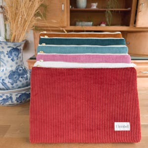 Puede incluir: Una pila de neceseres con cremallera de pana en varios colores, incluyendo rojo, blanco, rosa, turquesa y marrón. Cada neceser tiene una cremallera blanca y una pequeña etiqueta blanca con el nombre de la marca.