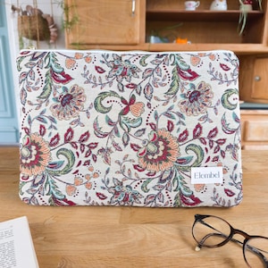 Può includere: Pochette rettangolare con motivo floreale su sfondo bianco, con fiori e foglie rosse, verdi e arancioni. La pochette ha una cerniera dorata e una piccola etichetta bianca con la scritta "Elembel".
