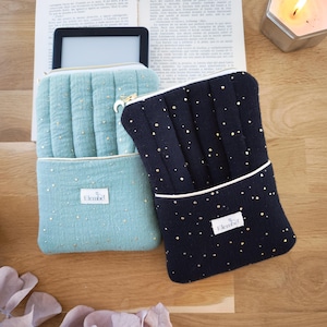 Puede incluir: Dos fundas acolchadas para lectores electrónicos. Una es azul claro con motas doradas y una cremallera dorada. La otra es azul marino con motas doradas y un ribete blanco. Ambas tienen una pequeña etiqueta blanca con el nombre de la marca.