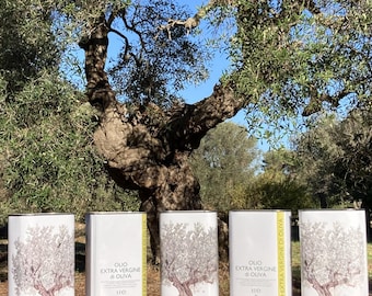 May include: Five white rectangular olive oil tins with a tree illustration and the text "OLIO EXTRA VERGINE DI OLIVA". The tins are arranged on a wooden surface, with a large olive tree in the background.