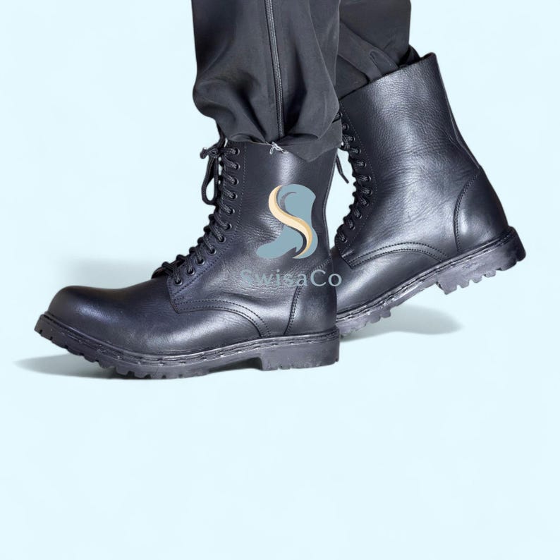 Pode incluir: Botas de combate de couro preto com design de atacadores. As botas t&ecirc;m sola grossa e s&atilde;o usadas com cal&ccedil;as pretas. O texto "SwisaCo" &eacute; vis&iacute;vel na lateral das botas.