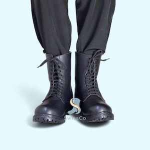 Pode incluir: Botas de combate de couro preto com atacadores. As botas t&ecirc;m uma sola resistente e s&atilde;o usadas com cal&ccedil;as pretas. Estas botas s&atilde;o um estilo cl&aacute;ssico, adequado para v&aacute;rias roupas.