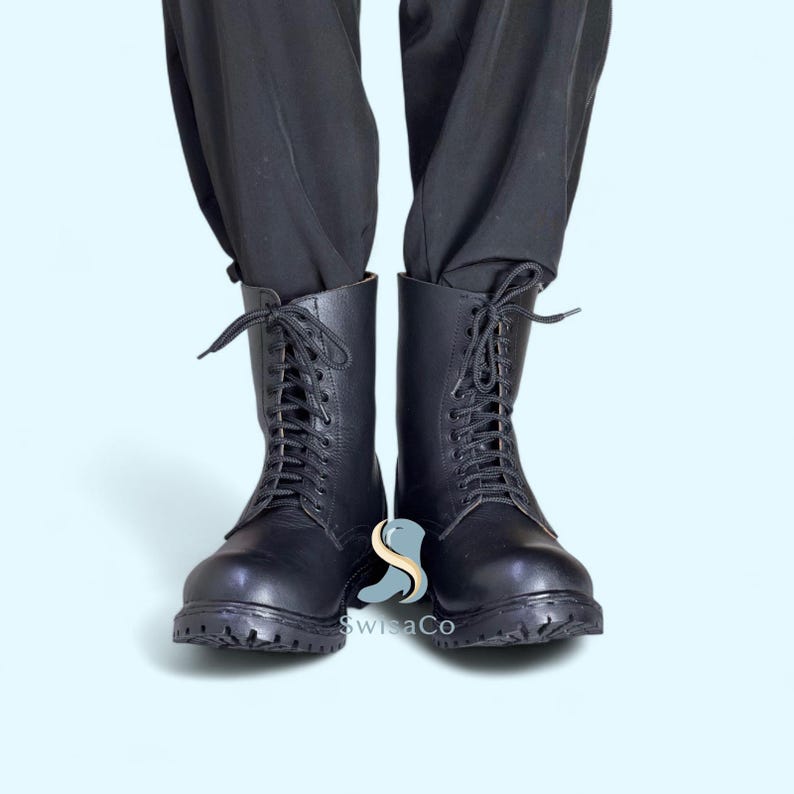 Pode incluir: Botas de combate de couro preto com atacadores. As botas t&ecirc;m uma sola resistente e s&atilde;o usadas com cal&ccedil;as pretas. Estas botas s&atilde;o um estilo cl&aacute;ssico, adequado para v&aacute;rias roupas.