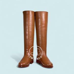 Puede incluir: Un par de botas altas de cuero marrón. Las botas tienen un acabado liso y pulido y un diseño clásico. Las suelas son de un tono marrón más oscuro. Las botas se muestran sobre un fondo azul claro.