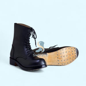 Puede incluir: Un par de botas de cuero negro con cordones, caña alta y cordones negros. Una bota está de pie mientras que la otra está de lado, revelando una suela de cuero color canela claro reforzada con numerosos clavos metálicos en toda la superficie.
