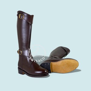 以下が含まれることがあります： 金色のバックルが付いた茶色の革製ライディングブーツ。片方のブーツは直立しており、もう片方は横に倒れていて、ソールが見えています。このブーツは、乗馬活動に適したクラシックなデザインです。